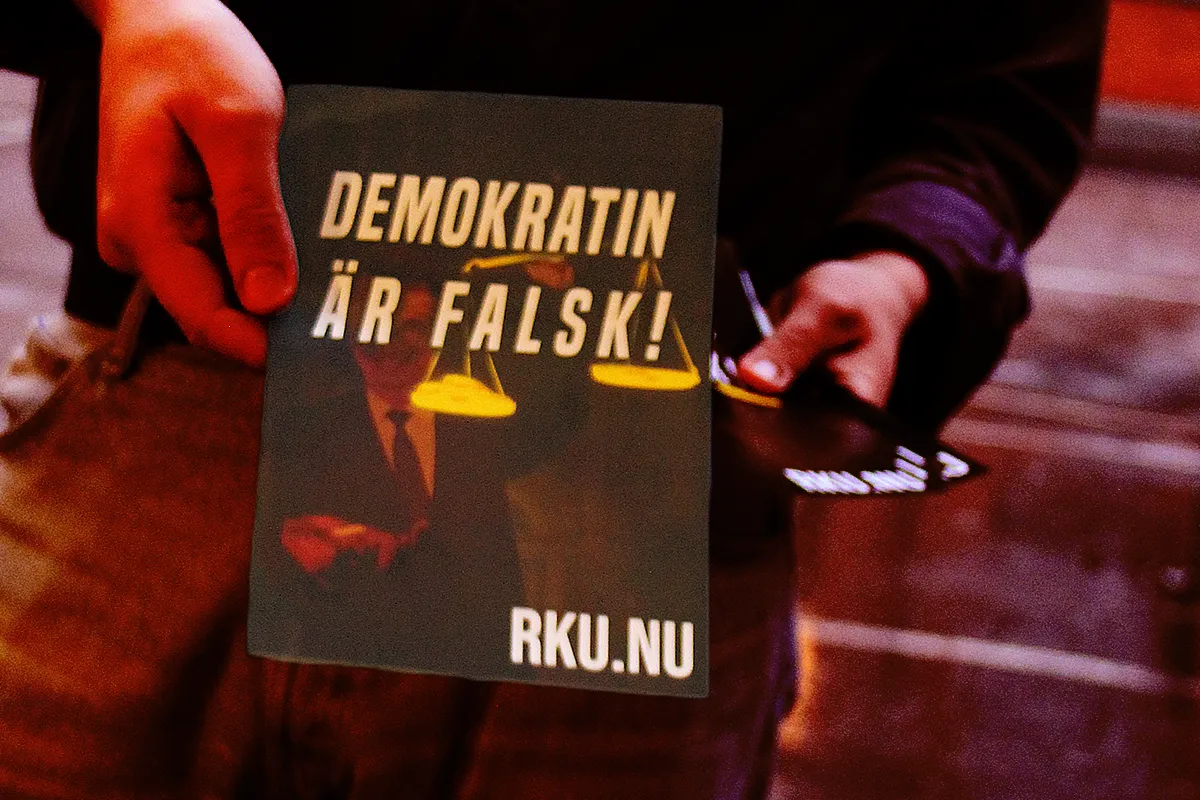 En person räcker över ett flygblad med texten "Demokratin är falsk!" på